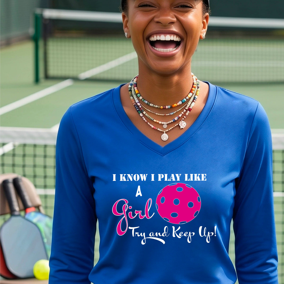 A woman laughing while wearing a royal blue long-sleeve v-neck  t-shirt with a pink and white graphic that reads 'I KNOW I PLAY LIKE A Girl Try and Keep Up!' with a pink pickleball illustration.