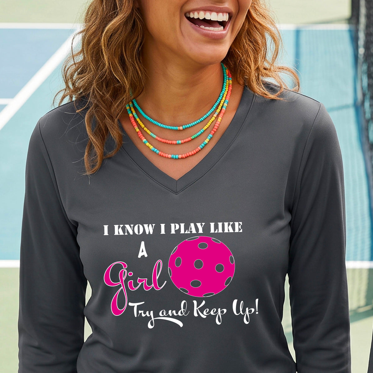A woman laughing while wearing a dark gray long-sleeve v-neck  t-shirt with a pink and white graphic that reads 'I KNOW I PLAY LIKE A Girl Try and Keep Up!' with a pink pickleball illustration.