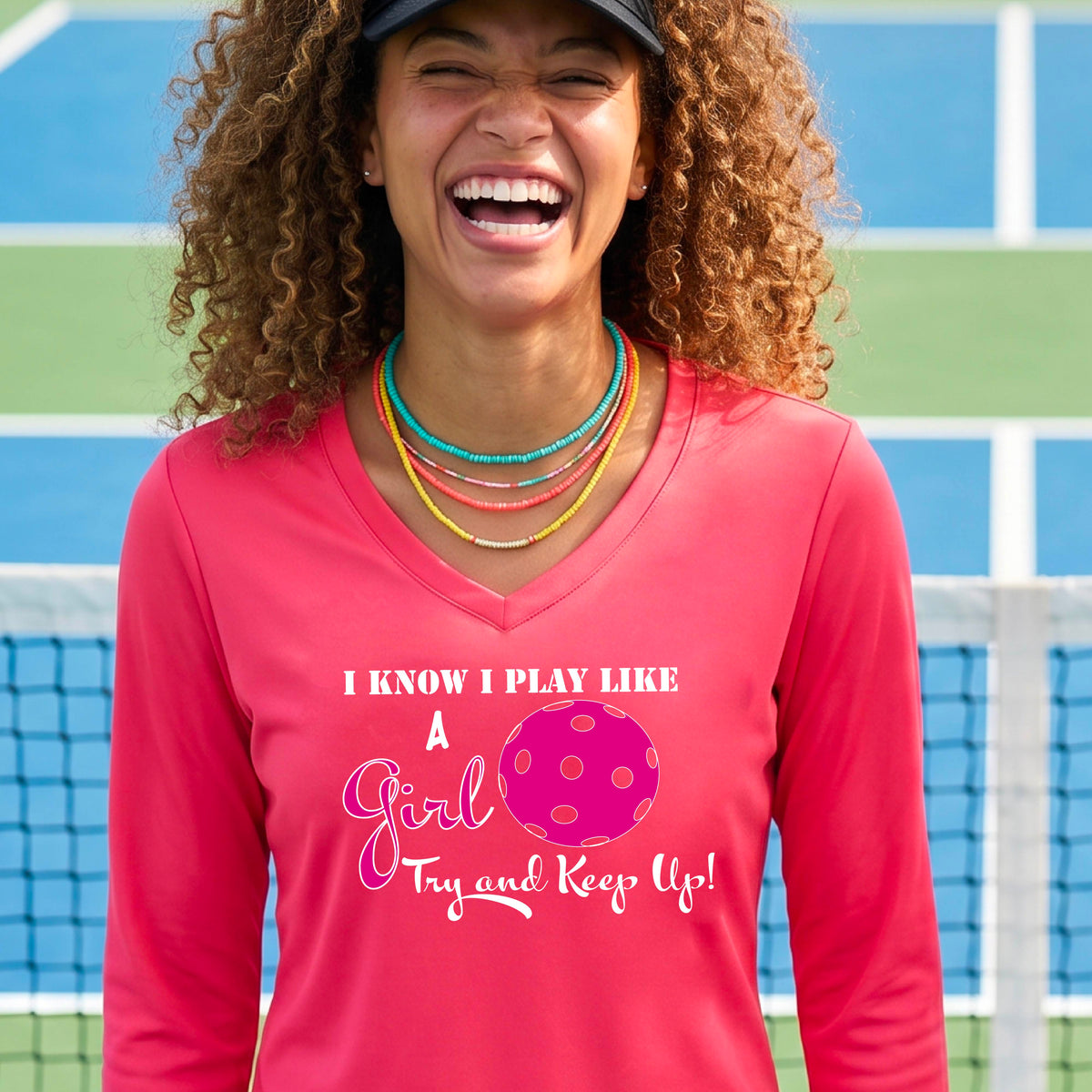 A woman laughing while wearing a bright coral colored long-sleeve v-neck  t-shirt with a pink and white graphic that reads 'I KNOW I PLAY LIKE A Girl Try and Keep Up!' with a pink pickleball illustration.