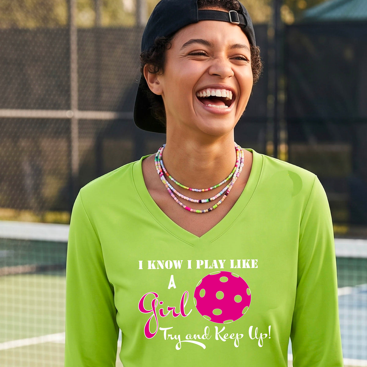 A woman laughing while wearing a lime green long-sleeve v-neck  t-shirt with a pink and white graphic that reads 'I KNOW I PLAY LIKE A Girl Try and Keep Up!' with a pink pickleball illustration.