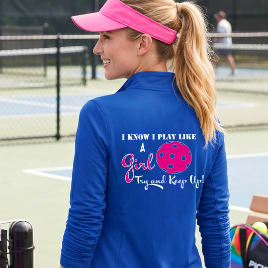 A woman laughing while wearing a royal blue quarter-zip shirt with a pink and white graphic that reads 'I KNOW I PLAY LIKE A Girl Try and Keep Up!' with a pink pickleball illustration.