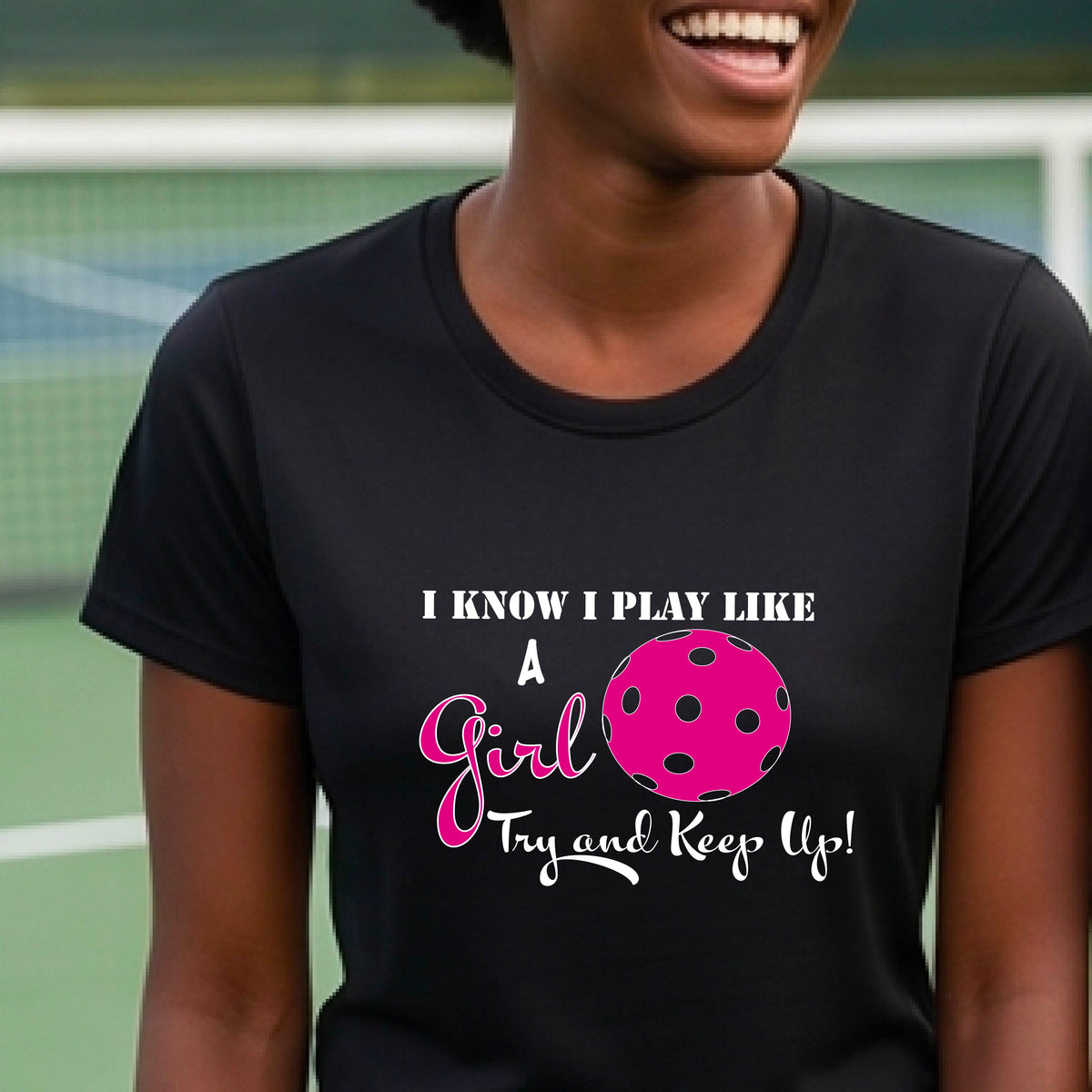 A woman laughing while wearing a black short-sleeve t-shirt with a pink and white graphic that reads 'I KNOW I PLAY LIKE A Girl Try and Keep Up!' with a pink pickleball illustration.