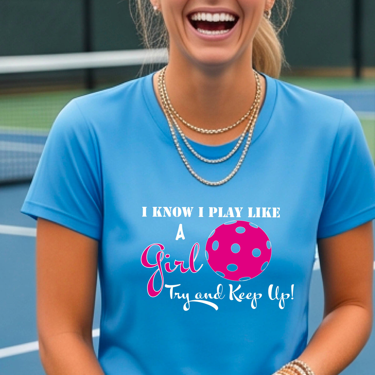 A woman laughing while wearing a blue short-sleeve t-shirt with a pink and white graphic that reads 'I KNOW I PLAY LIKE A Girl Try and Keep Up!' with a pink pickleball illustration.