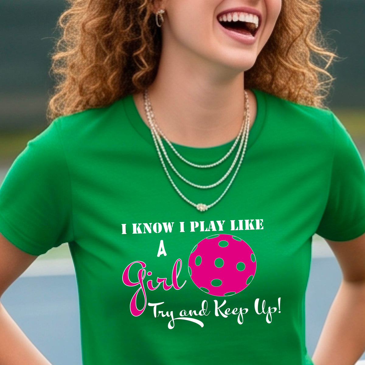 A woman laughing while wearing a green short-sleeve t-shirt with a pink and white graphic that reads 'I KNOW I PLAY LIKE A Girl Try and Keep Up!' with a pink pickleball illustration.