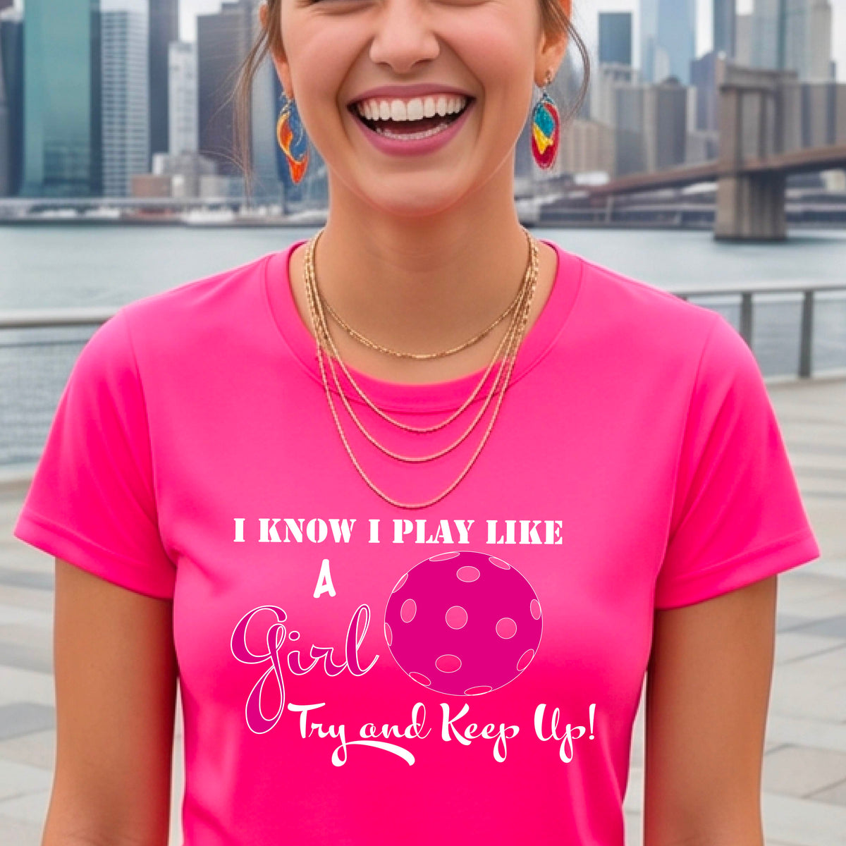 A woman laughing while wearing a pink short-sleeve t-shirt with a pink and white graphic that reads 'I KNOW I PLAY LIKE A Girl Try and Keep Up!' with a pink pickleball illustration.