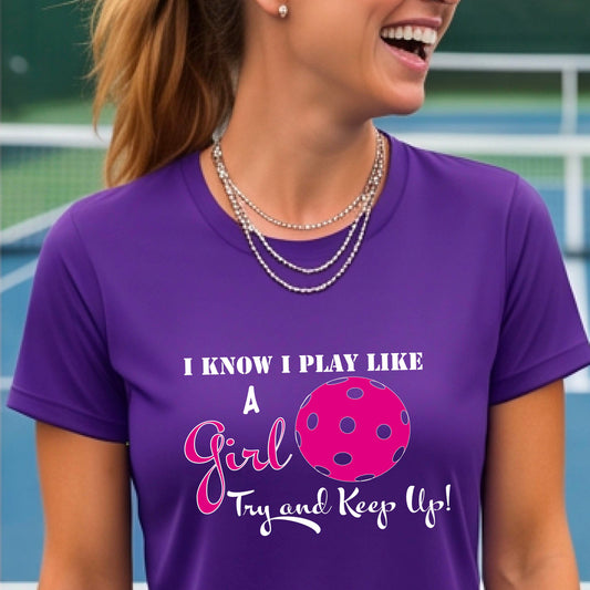 A woman laughing while wearing a purple short-sleeve t-shirt with a pink and white graphic that reads 'I KNOW I PLAY LIKE A Girl Try and Keep Up!' with a pink pickleball illustration.