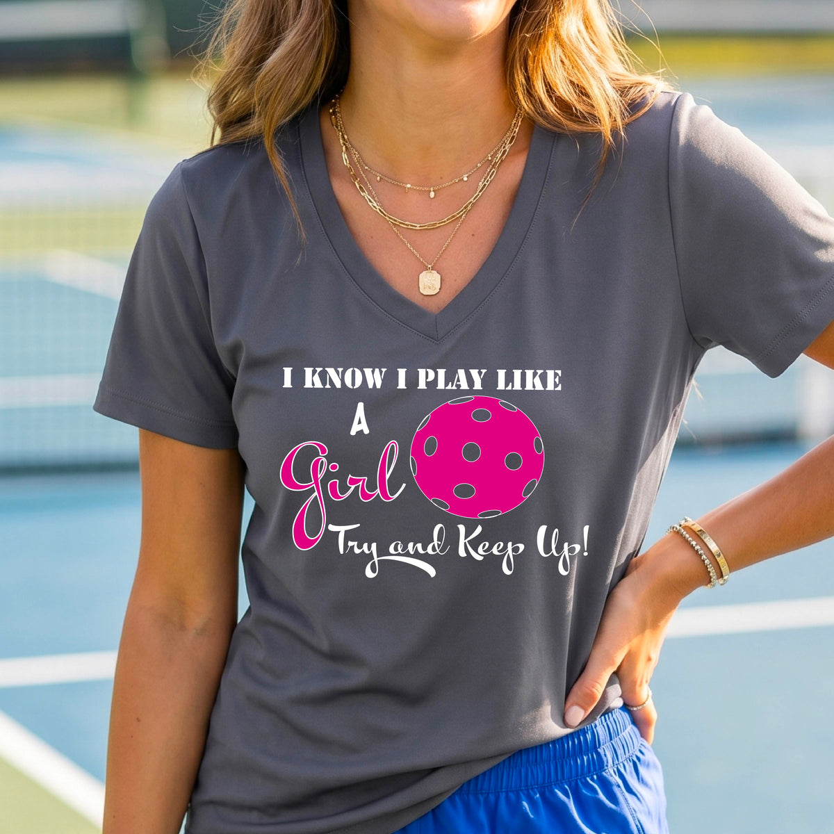 A woman laughing while wearing a dark gray short-sleeve v-neck  t-shirt with a pink and white graphic that reads 'I KNOW I PLAY LIKE A Girl Try and Keep Up!' with a pink pickleball illustration.
