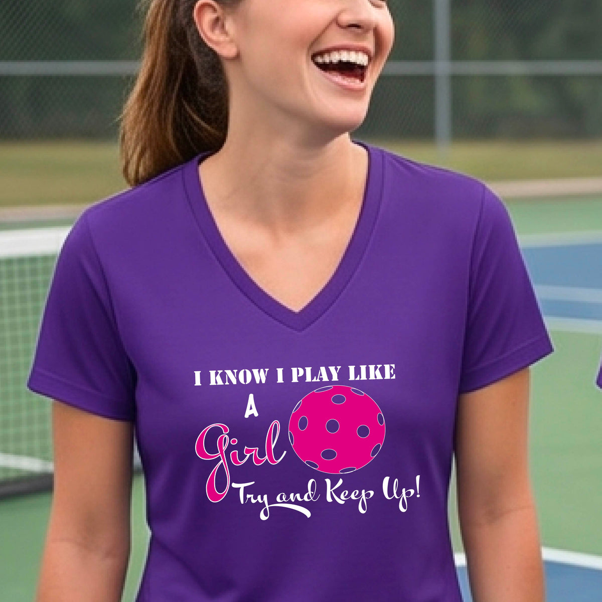 A woman laughing while wearing a purple short-sleeve v-neck  t-shirt with a pink and white graphic that reads 'I KNOW I PLAY LIKE A Girl Try and Keep Up!' with a pink pickleball illustration.