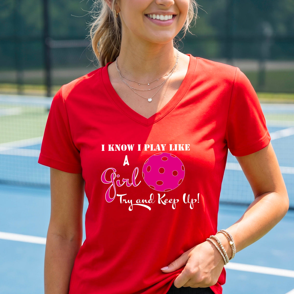 A woman laughing while wearing a red short-sleeve v-neck  t-shirt with a pink and white graphic that reads 'I KNOW I PLAY LIKE A Girl Try and Keep Up!' with a pink pickleball illustration.