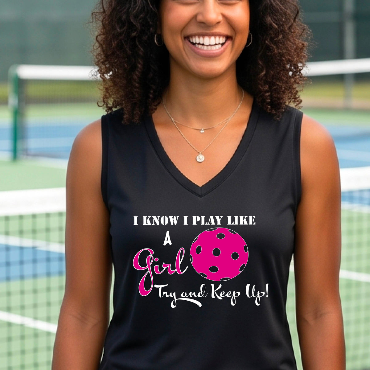 A close-up of a woman wearing a black sleeveless with a white graphic of ‘I KNOW I PLAY LIKE A GIRL TRY AND KEEP UP’ in white and pink with a pink pickleball icon, in front of a pickleball court. 