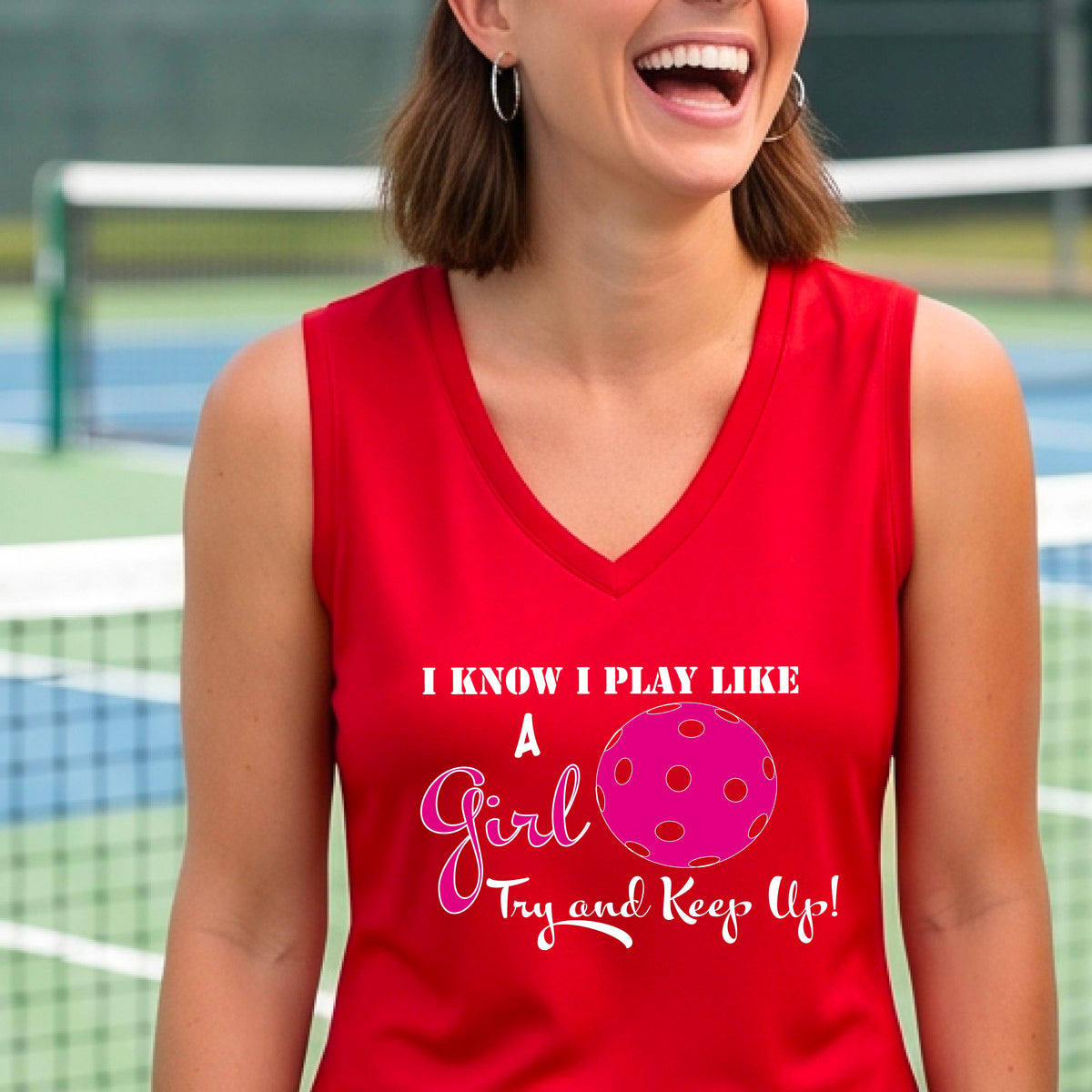 A close-up of a woman wearing a red sleeveless with a white graphic of ‘I KNOW I PLAY LIKE A GIRL TRY AND KEEP UP’ in white and pink with a pink pickleball icon, in front of a pickleball court. 