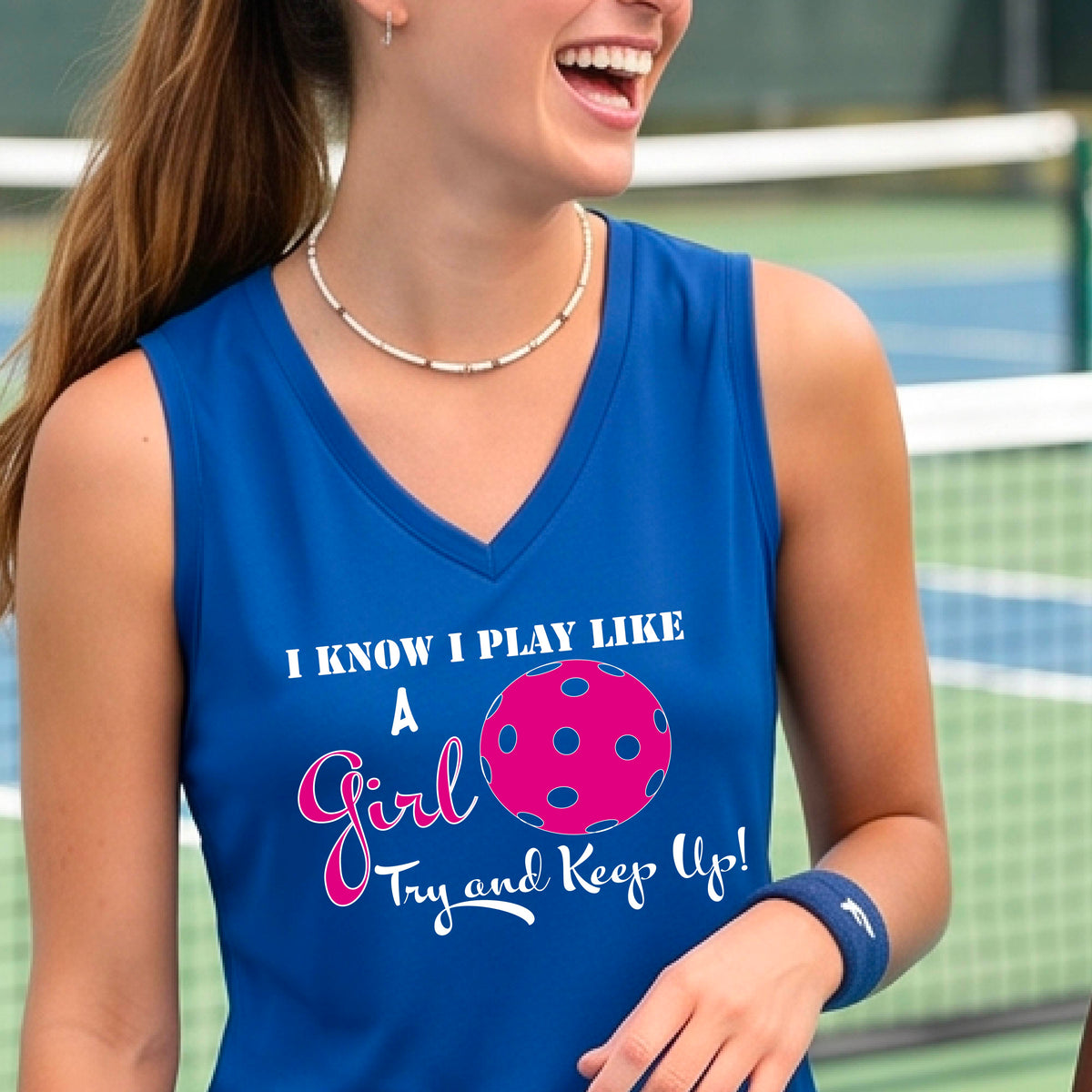 A close-up of a woman wearing a royal blue sleeveless with a white graphic of ‘I KNOW I PLAY LIKE A GIRL TRY AND KEEP UP’ in white and pink with a pink pickleball icon, in front of a pickleball court. 