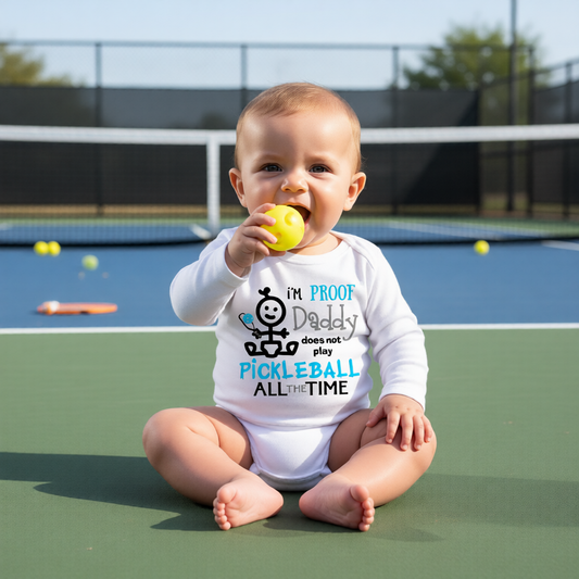 A baby sitting on a green pickleball court wearing a white long-sleeve bodysuit with a graphic that reads 'I'M PROOF Daddy does not play PICKLEBALL ALL THE TIME' in black and blue text. 