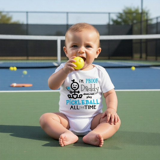 A baby sitting on a pickleball court wearing a white graphic t-shirt that reads 'I'M PROOF Daddy does not play PICKLEBALL ALL THE TIME' with a cartoon character. 