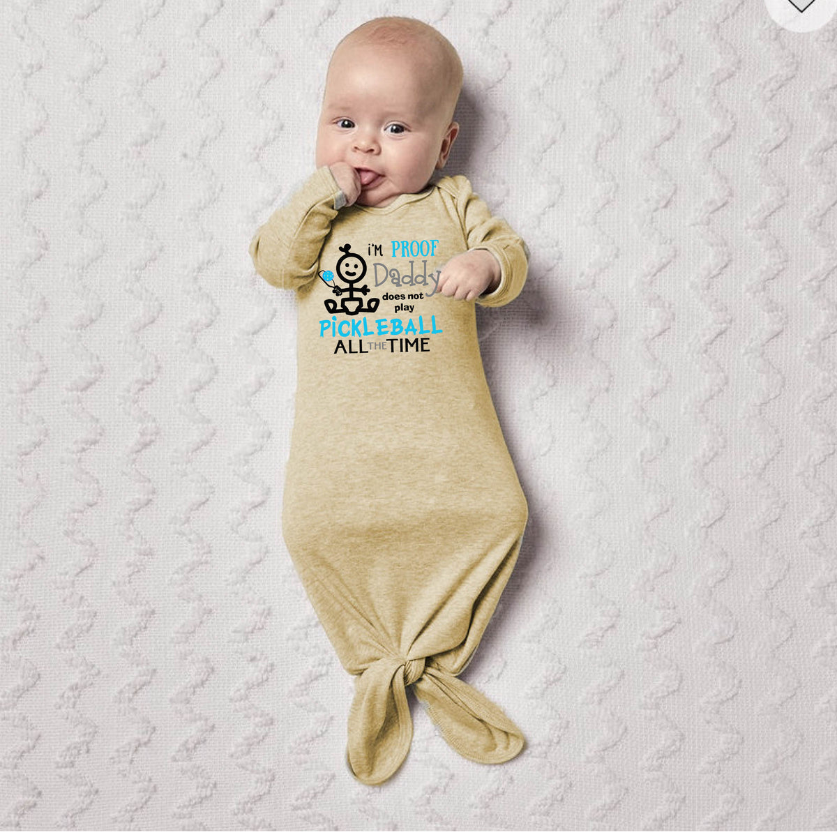 A baby wearing a beige long-sleeve knotted infant gown, with a graphic print that reads 'I'M PROOF Daddy Does not play PICKLEBALL ALL TIME' with a pickleball icon.