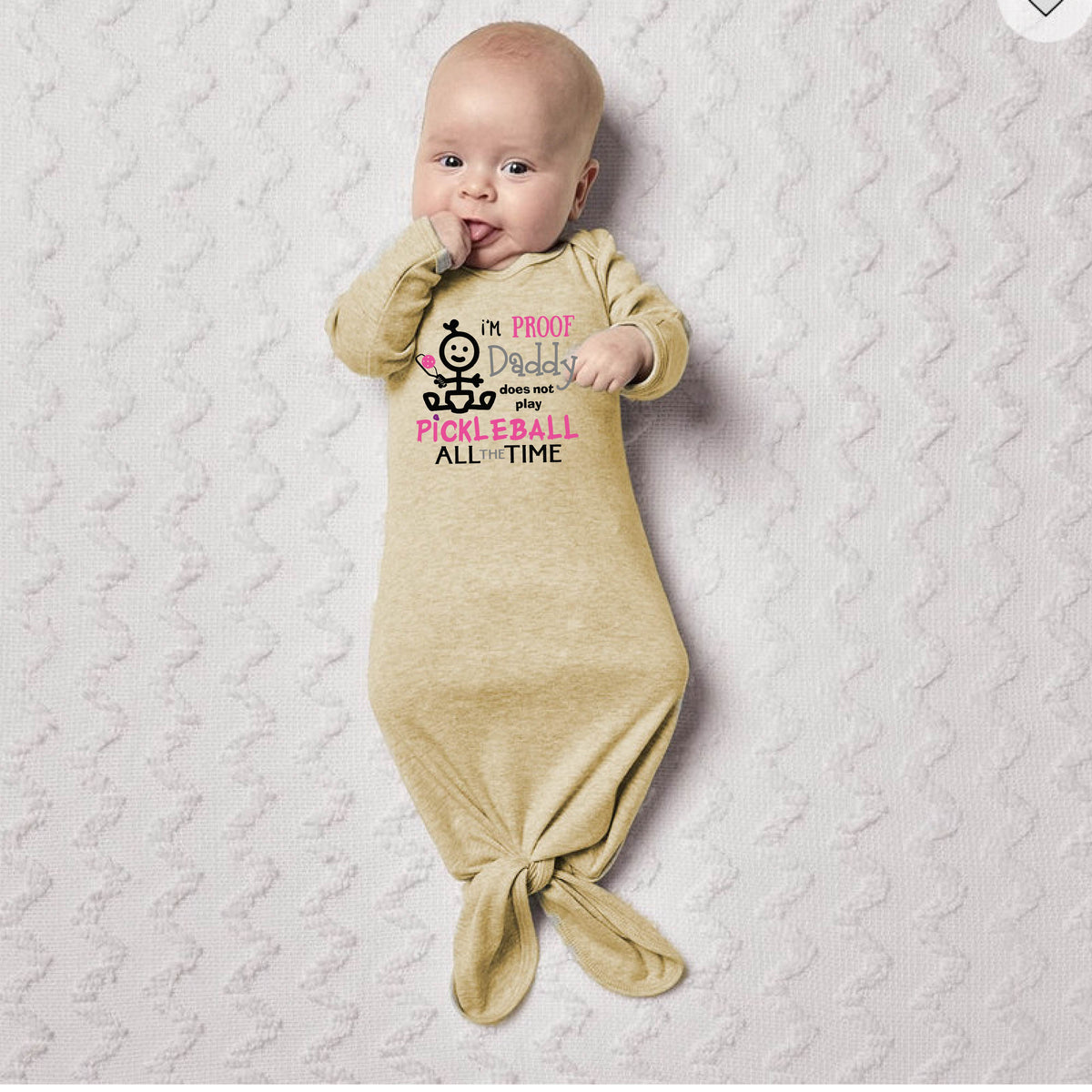 A baby wearing a beige long-sleeve knotted infant gown, with a graphic print that reads 'I'M PROOF Daddy Does not play PICKLEBALL ALL TIME' with a pickleball icon.