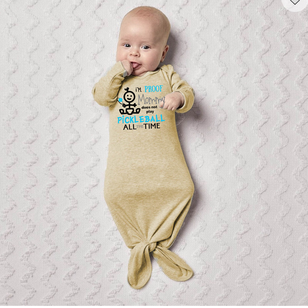 A baby wearing a beige long-sleeve knotted infant gown, with a graphic print that reads 'I'M PROOF Mommy Does not play PICKLEBALL ALL TIME' with a pickleball icon.