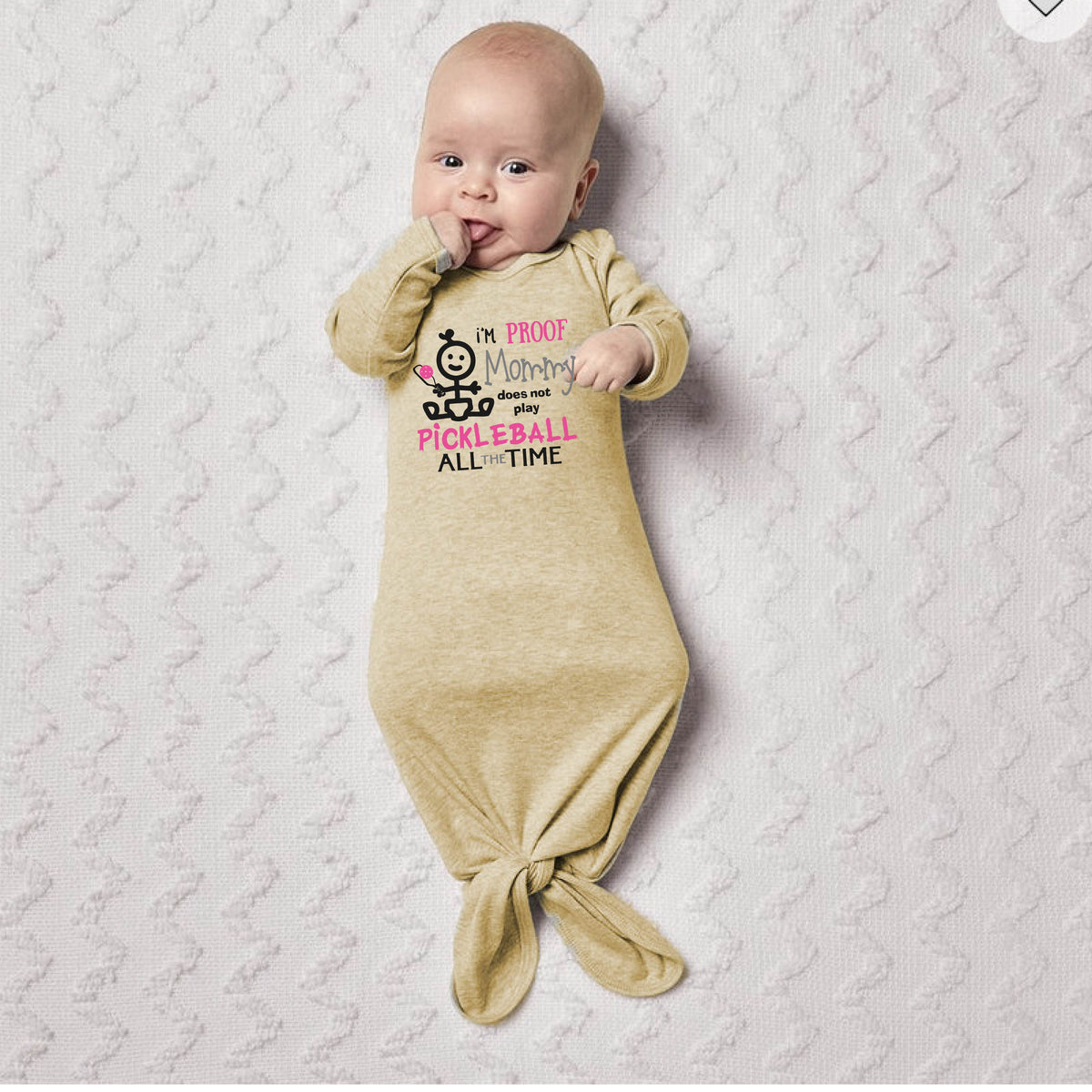 A baby wearing a beige long-sleeve knotted infant gown, with a graphic print that reads 'I'M PROOF Mommy Does not play PICKLEBALL ALL TIME' with a pickleball icon.