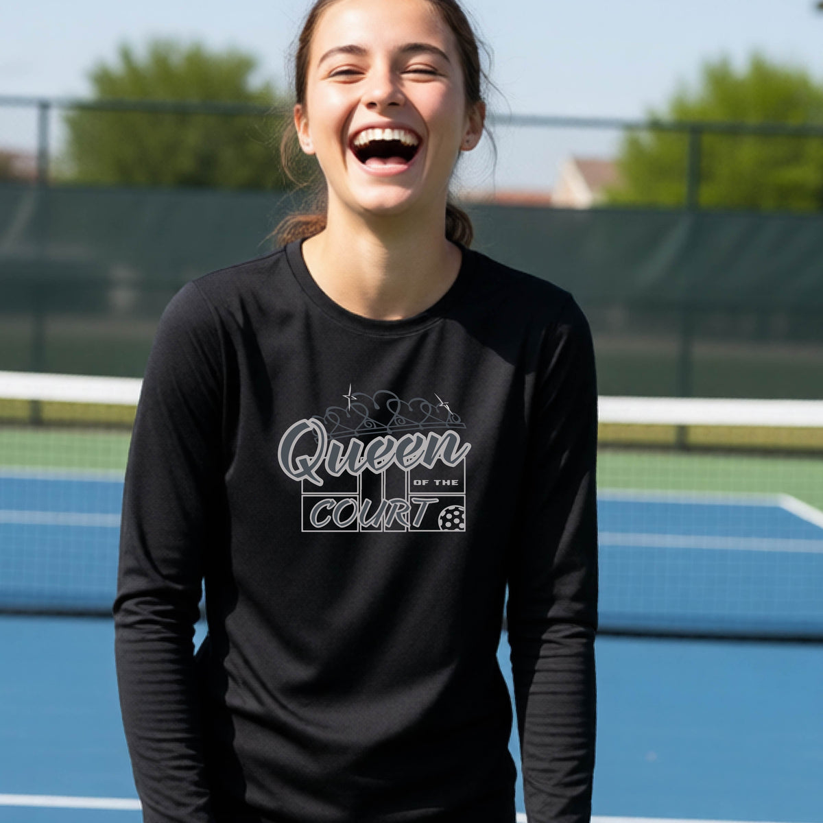 A smiling young woman stands on a pickleball court wearing a black long-sleeve shirt with a 'Queen of the Court' gray and white  graphic on the front.