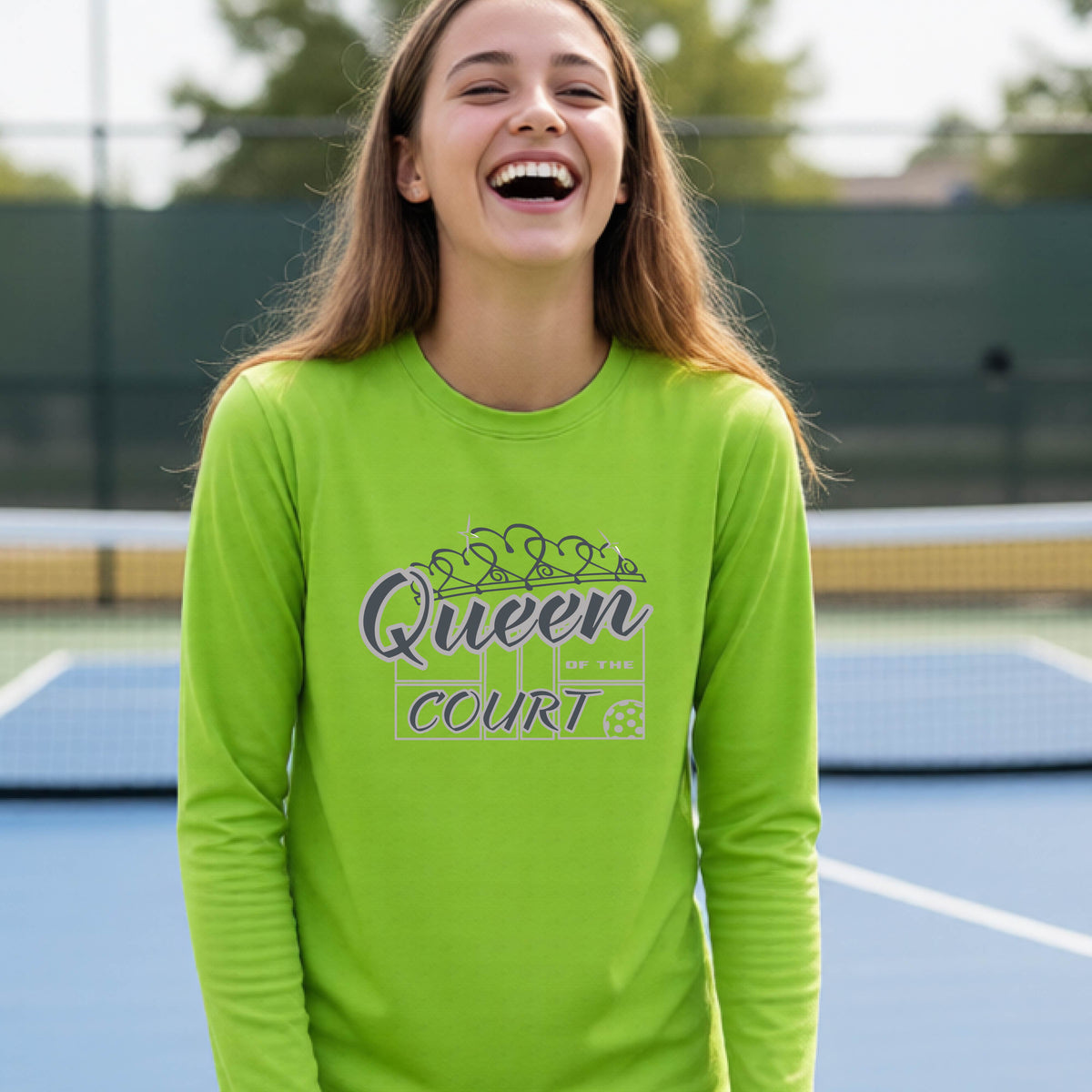 A smiling young woman stands on a pickleball court wearing a bright lime green long-sleeve shirt with a 'Queen of the Court' gray and white  graphic on the front.
