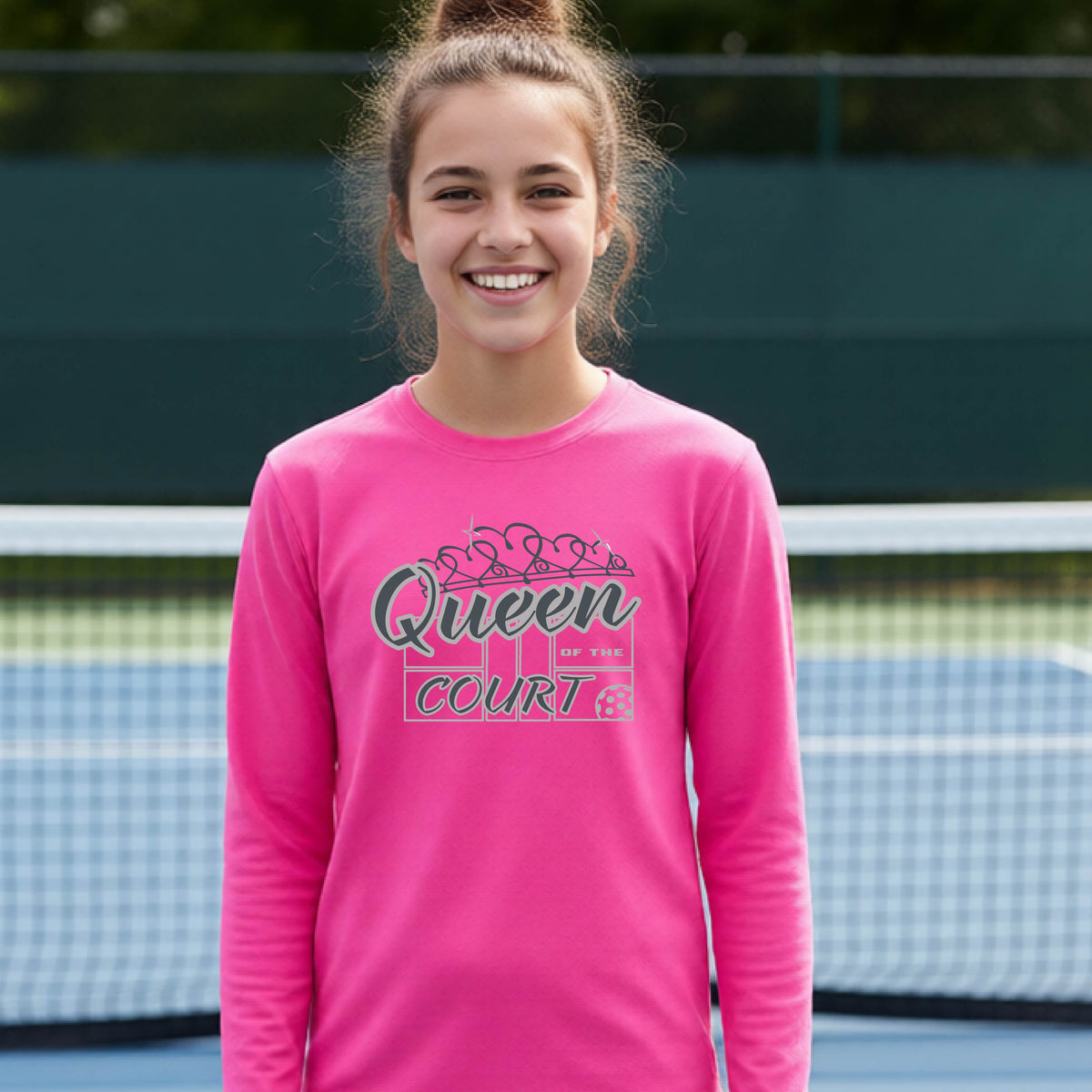 A smiling young woman stands on a pickleball court wearing a bright pink long-sleeve shirt with a 'Queen of the Court' gray and white  graphic on the front.