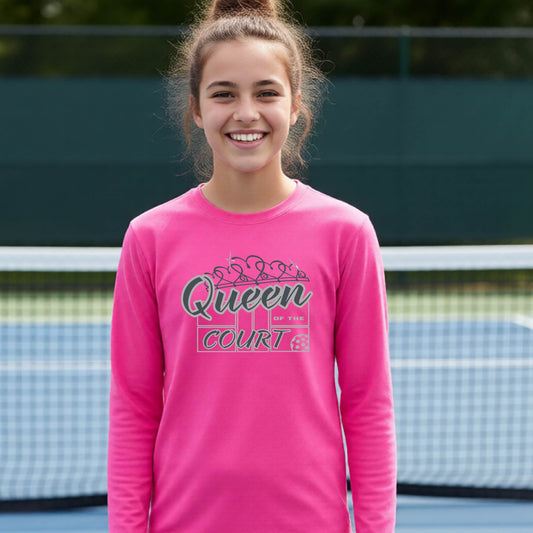 A smiling young woman stands on a pickleball court wearing a bright pink long-sleeve shirt with a 'Queen of the Court' gray and white  graphic on the front.