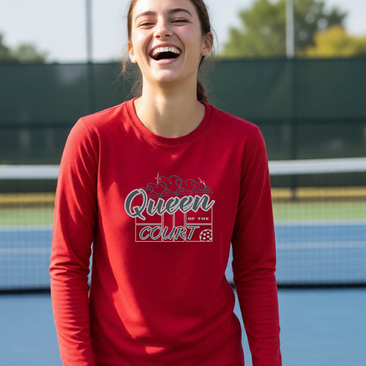 A smiling young woman stands on a pickleball court wearing a red long-sleeve shirt with a 'Queen of the Court' gray and white  graphic on the front.