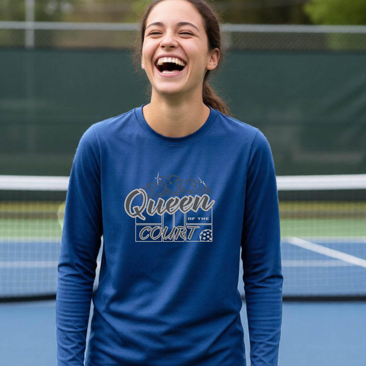 A smiling young woman stands on a pickleball court wearing a royal blue long-sleeve shirt with a 'Queen of the Court' gray and white  graphic on the front.