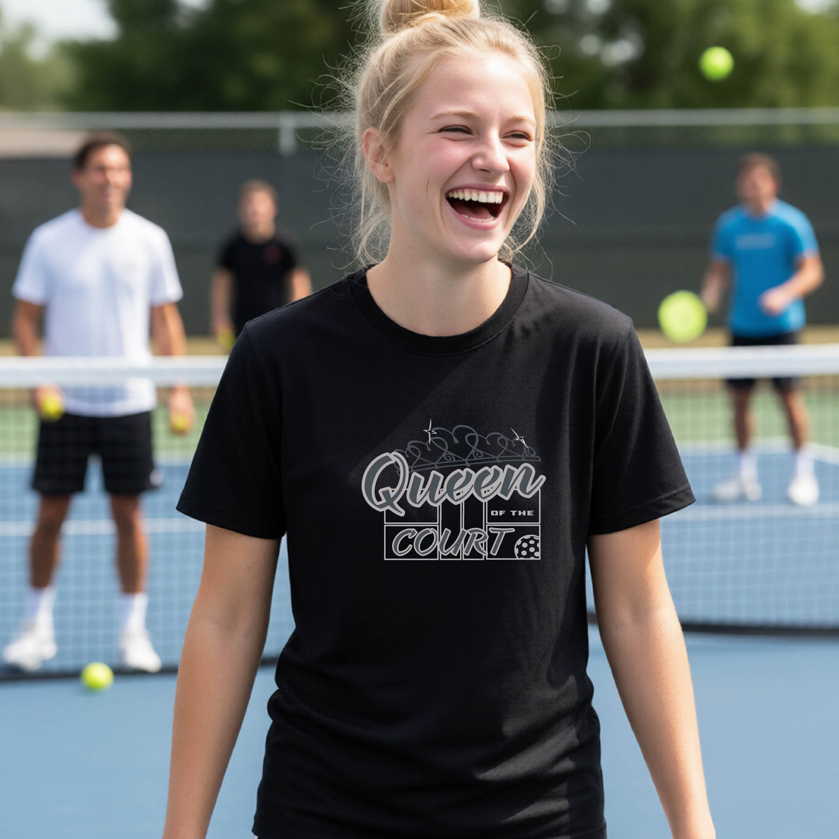 A young girl laughing on a blue pickleball court, wearing a black t-shirt with a graphic that reads 'Queen of the  Court' in gray and white.