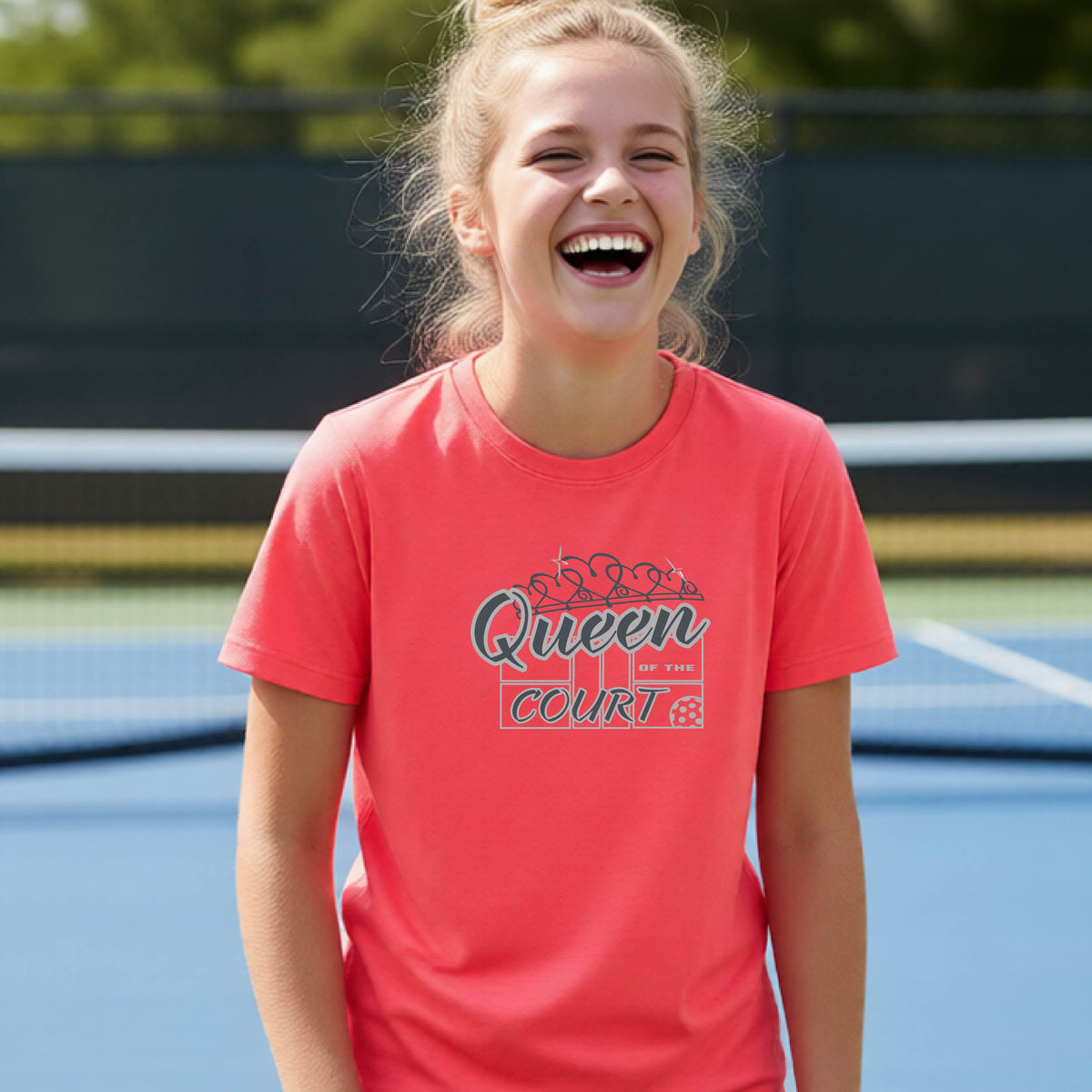 A young girl laughing on a blue pickleball court, wearing a coral pink t-shirt with a graphic that reads 'Queen of the  Court' in gray and white.