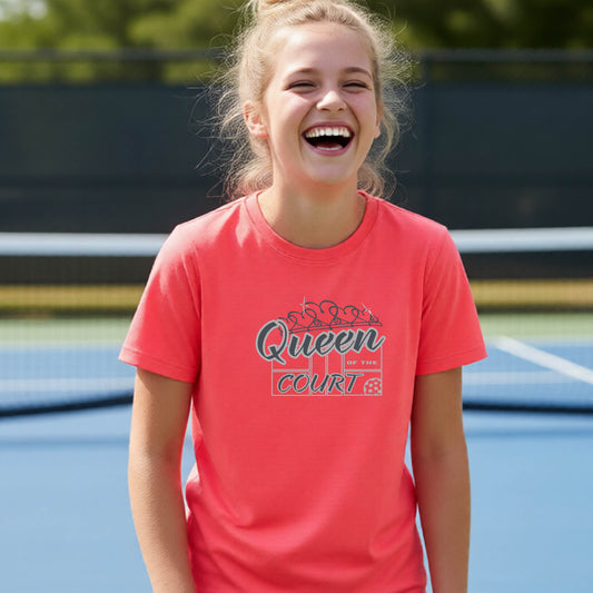 A young girl laughing on a blue pickleball court, wearing a coral pink t-shirt with a graphic that reads 'Queen of the  Court' in gray and white.