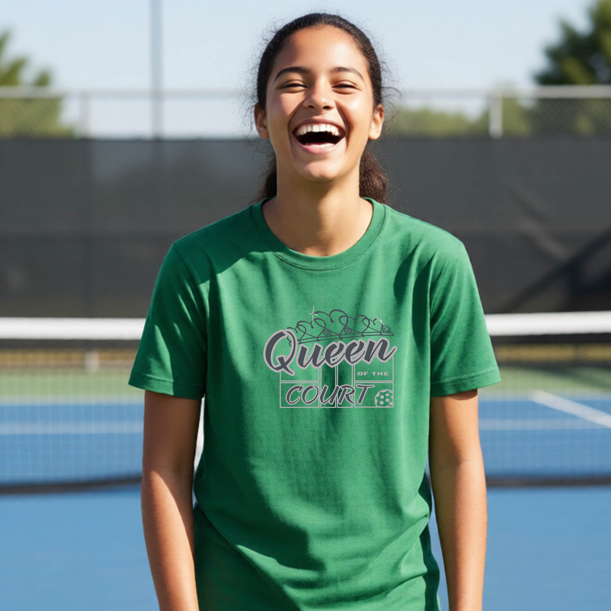 A young girl laughing on a blue pickleball court, wearing a green t-shirt with a graphic that reads 'Queen of the  Court' in gray and white.