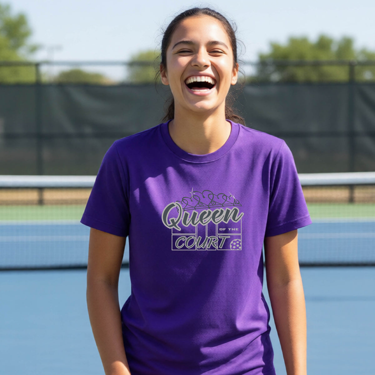 A young girl laughing on a blue pickleball court, wearing a purple t-shirt with a graphic that reads 'Queen of the  Court' in gray and white.