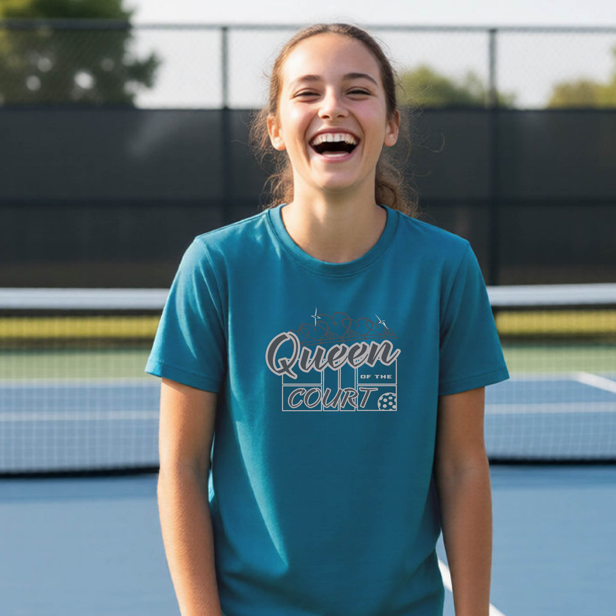 A young girl laughing on a blue pickleball court, wearing a teal t-shirt with a graphic that reads 'Queen of the  Court' in gray and white.
