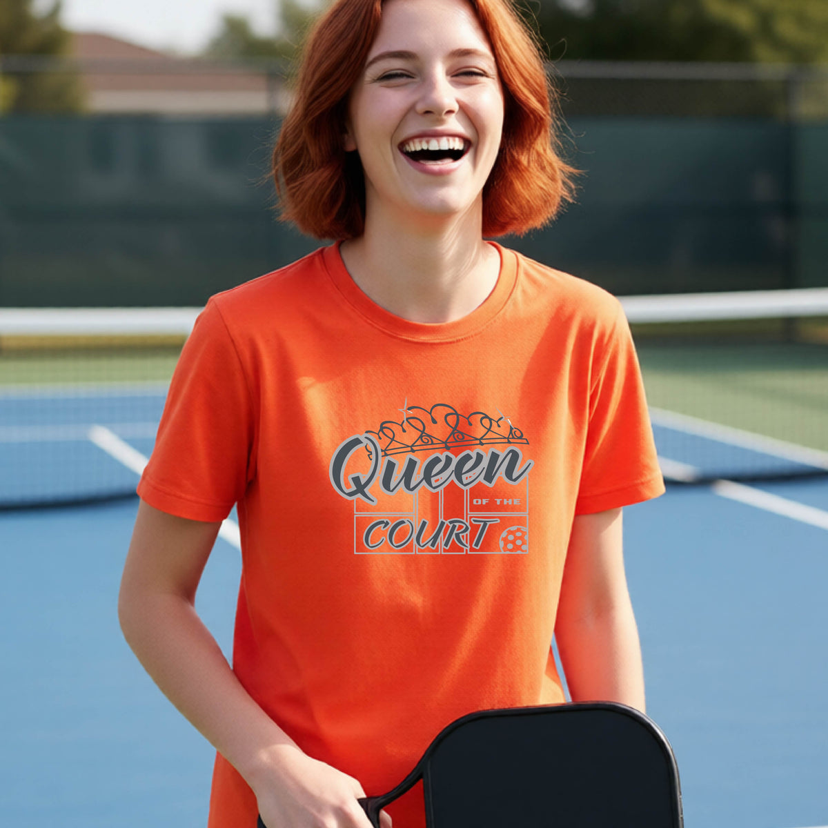A young girl laughing on a blue pickleball court, wearing an orange t-shirt with a graphic that reads 'Queen of the  Court' in gray and white.