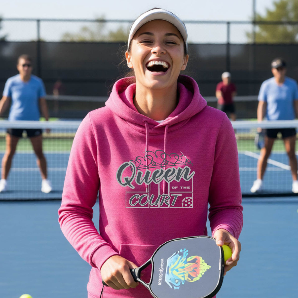 A woman laughing on a pickleball court wearing a pink hoodie with 'Queen of the Court' with a pickleball and court printed on the front,.