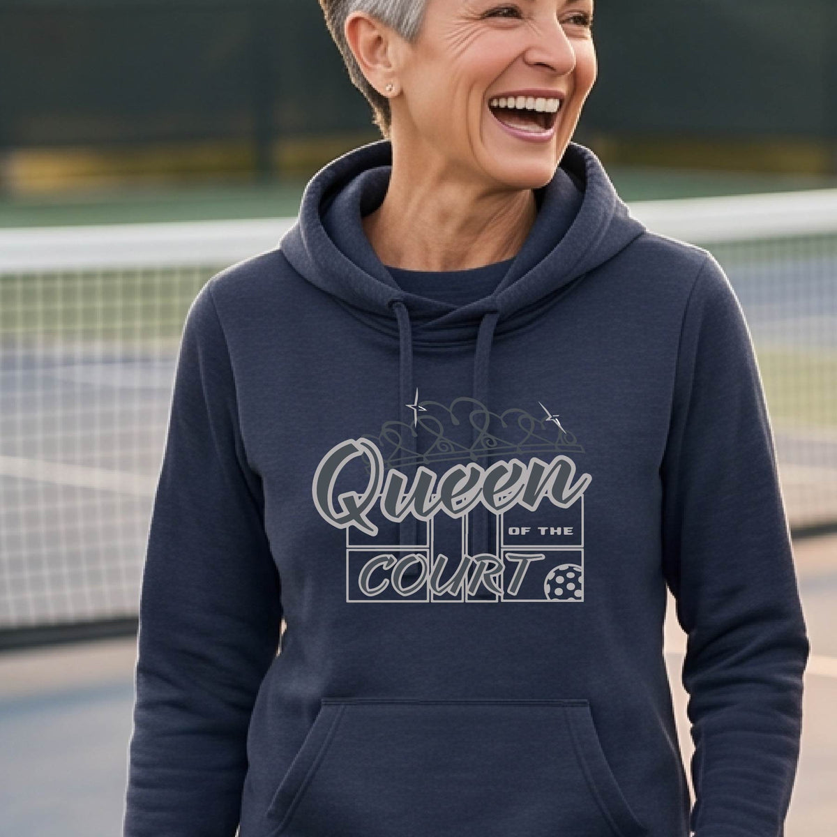 A woman laughing on a pickleball court wearing a navy blue hoodie with 'Queen of the Court' with a pickleball and court printed on the front,.