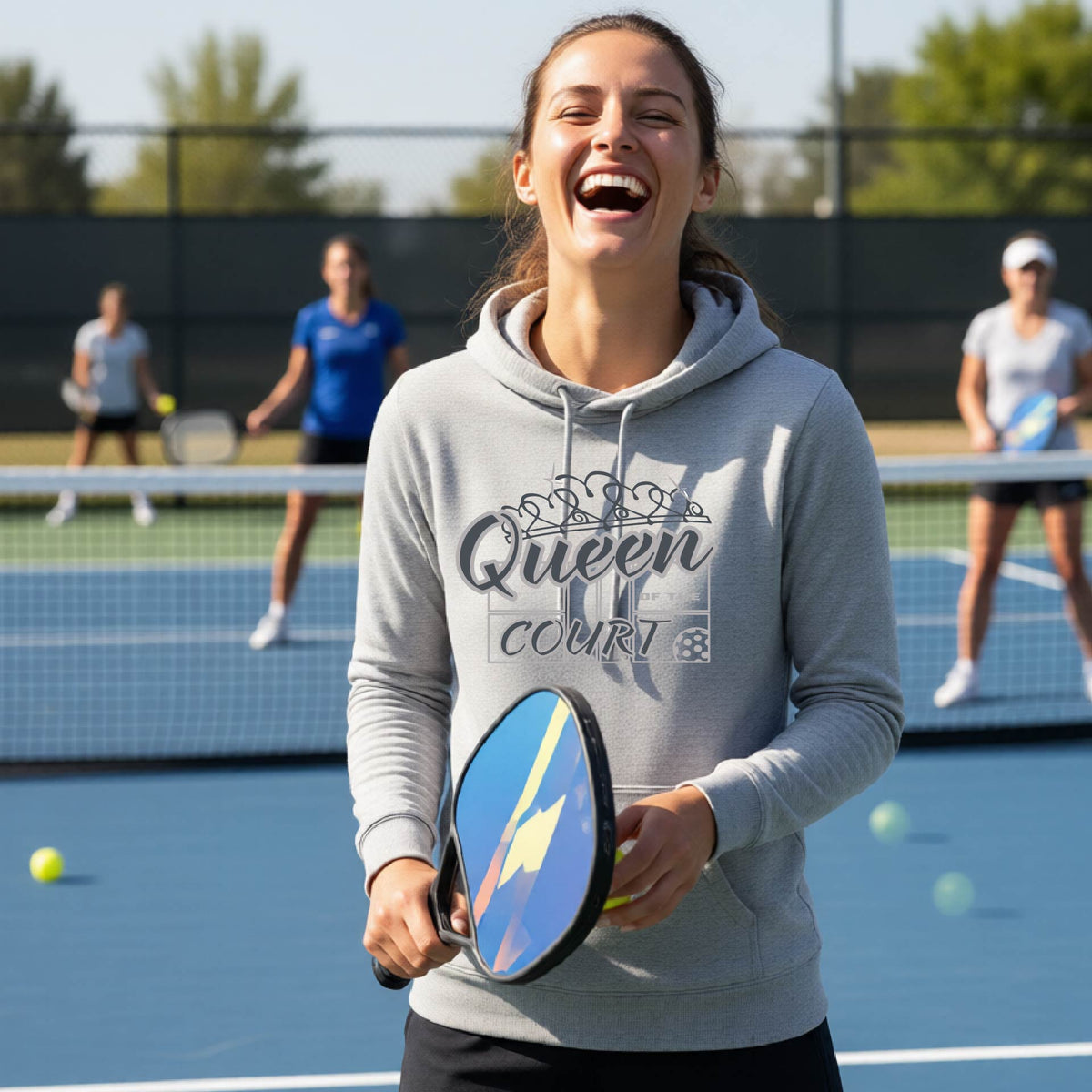 A woman laughing on a pickleball court wearing a light gray hoodie with 'Queen of the Court' with a pickleball and court printed on the front,.