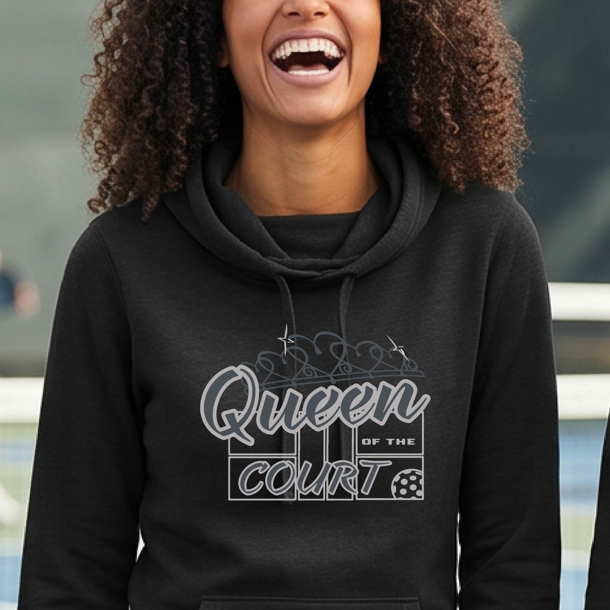 A woman laughing on a pickleball court wearing a black hoodie with 'Queen of the Court' with a pickleball and court printed on the front,.