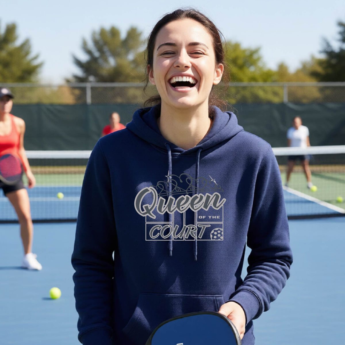 A woman laughing on an outdoor pickleball court wearing a navy blue hoodie with 'Queen of the Court' printed on the front. Other pickleball players are visible in the background.