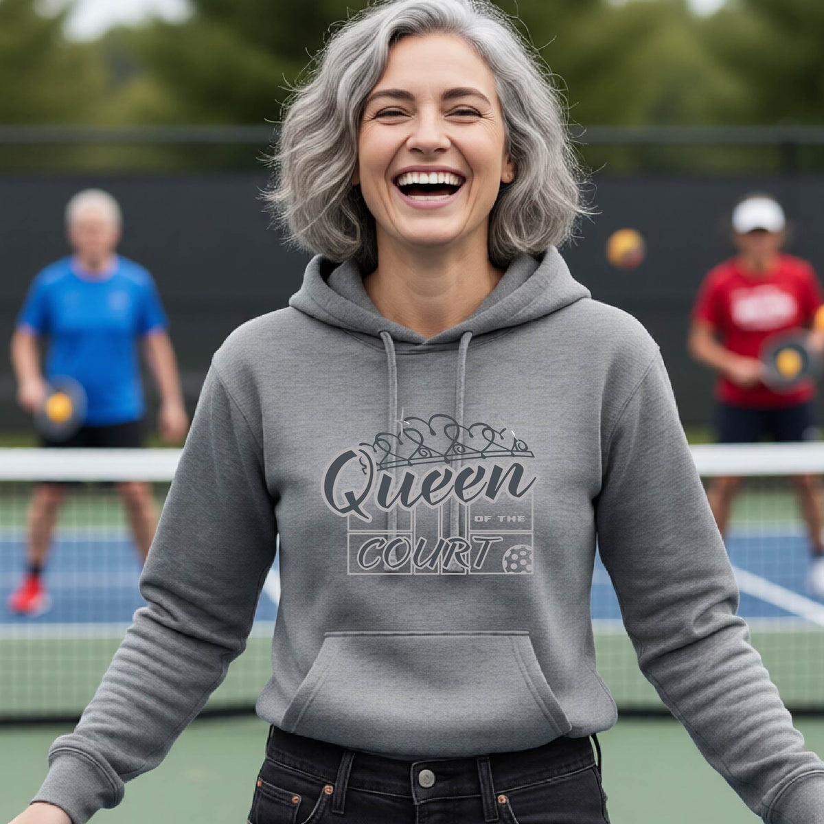 A woman laughing on an outdoor pickleball court wearing a gray hoodie with 'Queen of the Court' printed on the front. Other pickleball players are visible in the background.