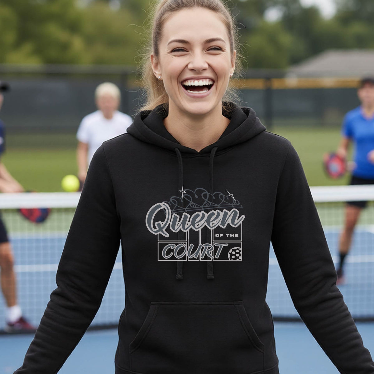 A woman laughing on an outdoor pickleball court wearing a black hoodie with 'Queen of the Court' printed on the front. Other pickleball players are visible in the background.