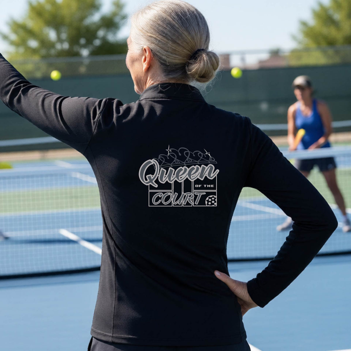 A woman laughing on a pickleball court, shown from behind, wearing a black quarter-zip shirt with a graphic that reads 'Queen of the Court' in gray and white.