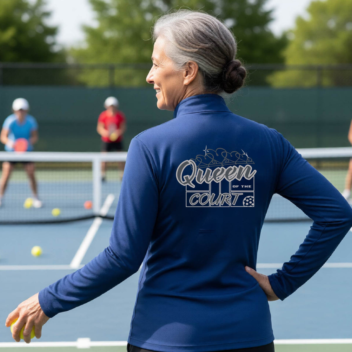 A woman laughing on a pickleball court, shown from behind, wearing a royal blue quarter-zip shirt with a graphic that reads 'Queen of the Court' in gray and white.