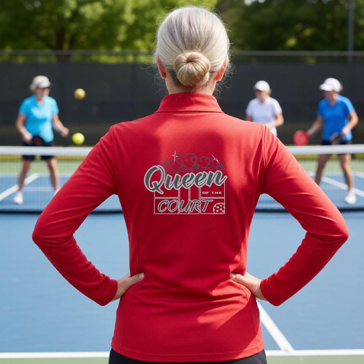 A woman laughing on a pickleball court, shown from behind, wearing a red quarter-zip shirt with a graphic that reads 'Queen of the Court' in gray and white.