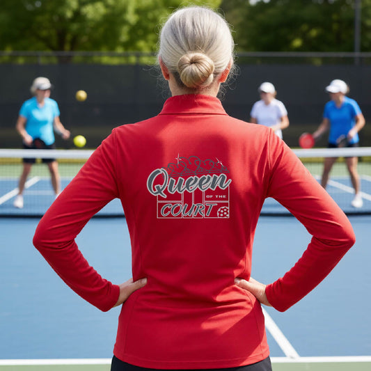A woman laughing on a pickleball court, shown from behind, wearing a red quarter-zip shirt with a graphic that reads 'Queen of the Court' in gray and white.