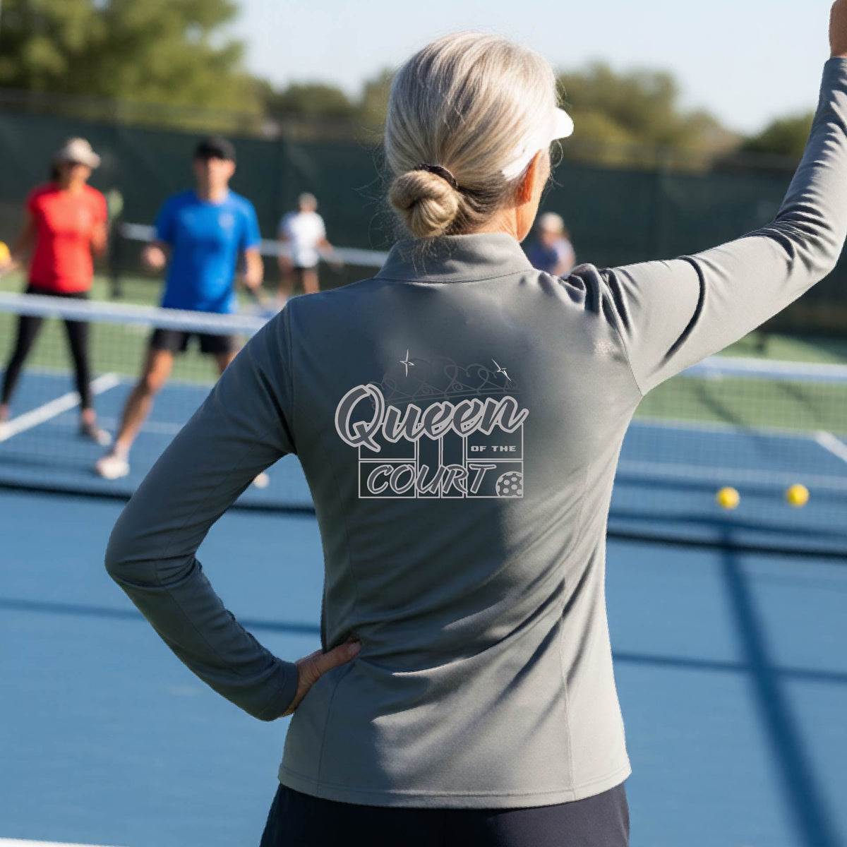 A woman laughing on a pickleball court, shown from behind, wearing a gray quarter-zip shirt with a graphic that reads 'Queen of the Court' in gray and white.