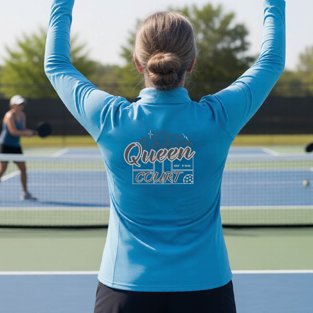 A woman laughing on a pickleball court, shown from behind, wearing a bright blue quarter-zip shirt with a graphic that reads 'Queen of the Court' in gray and white.