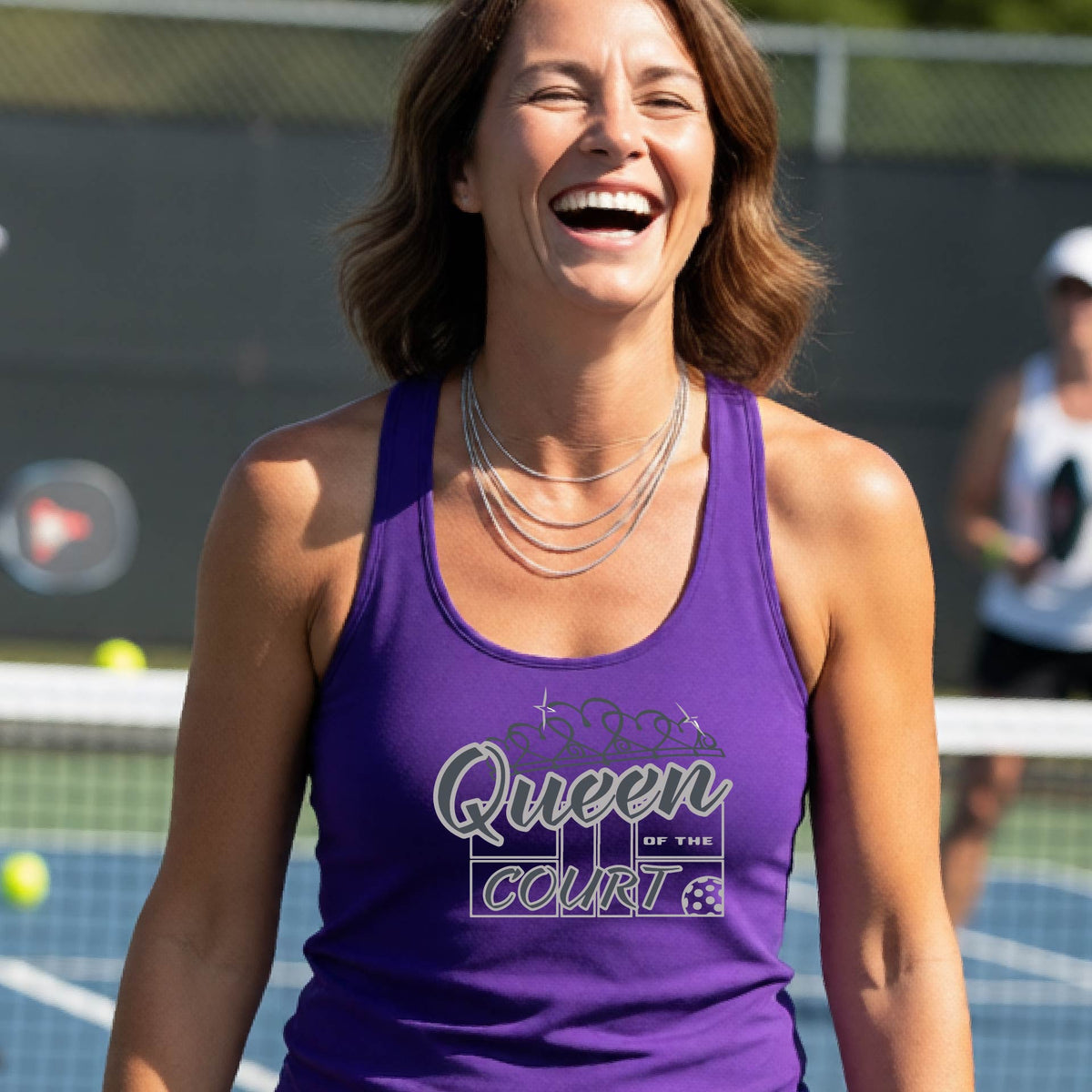 A woman laughing on a pickleball court, wearing a purple racerback tank top with a graphic that reads 'Queen of the Court' in gray and white and a pickleball graphic. 