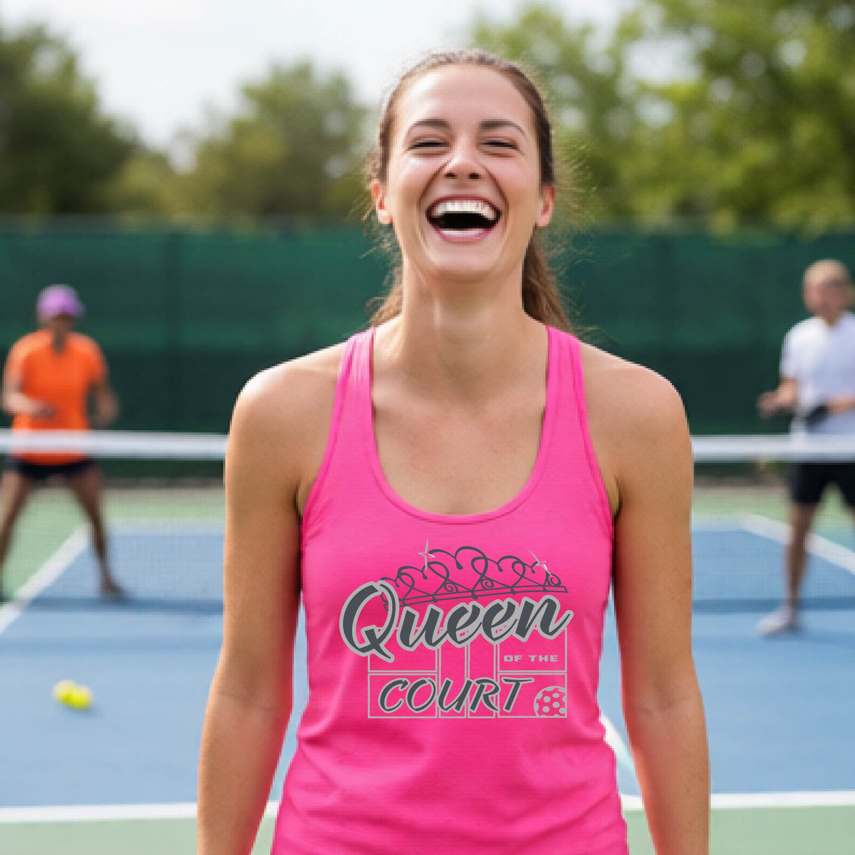 A woman laughing on a pickleball court, wearing a pink racerback tank top with a graphic that reads 'Queen of the Court' in gray and white and a pickleball graphic. 